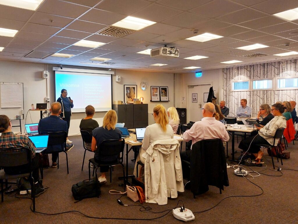 Rachel De Basso står framför en grupp med sittande människor och talar i mikrofon.